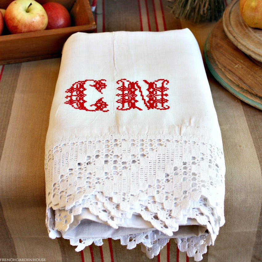 Antique French white linen sheet with red monogram and hand crochet lace edge on a table with apples in the background.