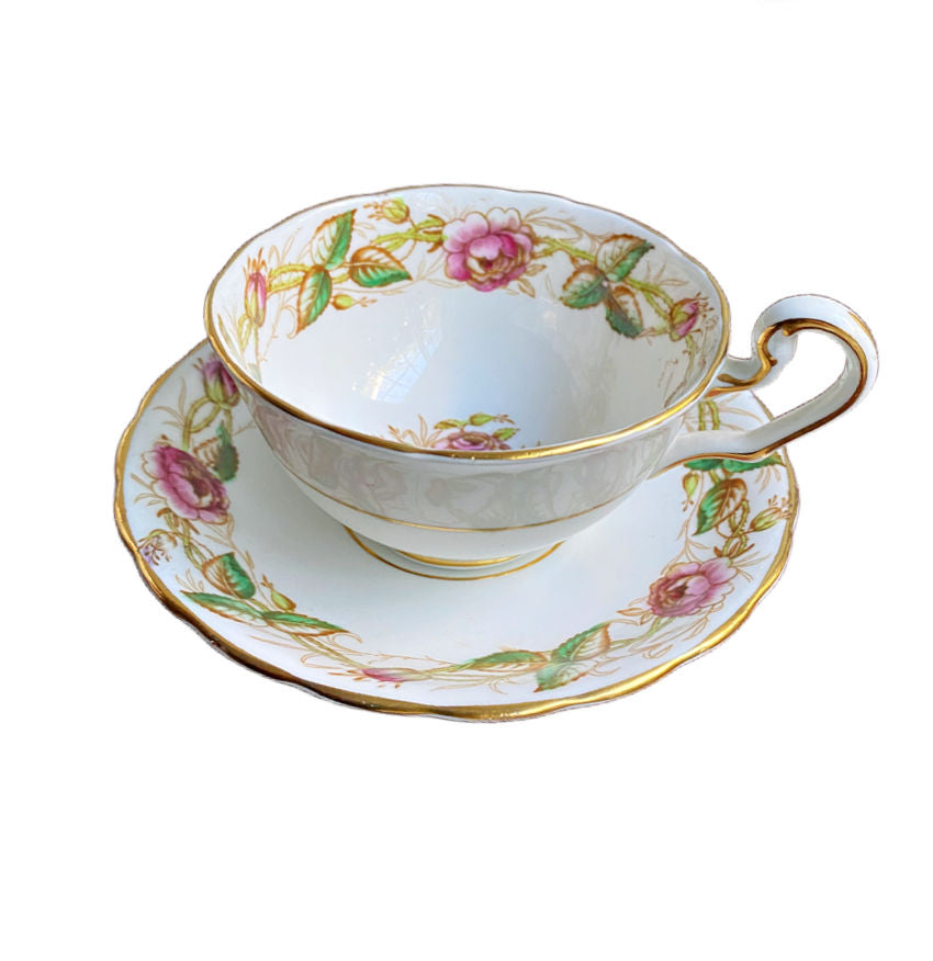 A hand painted English tea cup with pink roses and green leaves, accompanied by a saucer, against a white background.