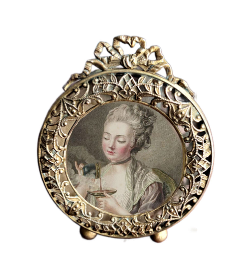 A 19th century Ormolu Gilt Bronze Picture Frame with a French ribbon bow at the top and a foliate design on the gilded brass oval frame, containing an image of a woman in a pastoral setting.