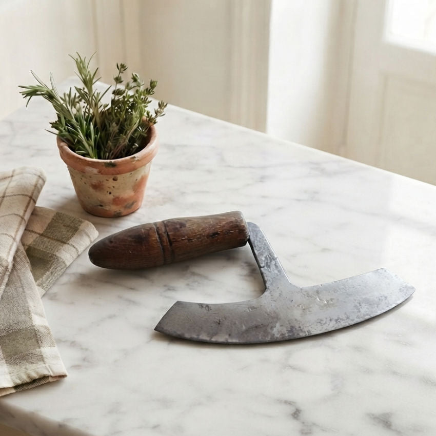 Antique 19th-century French hand-forged crescent blade chopper resting on a marble counter beside a linen towel and potted herbs