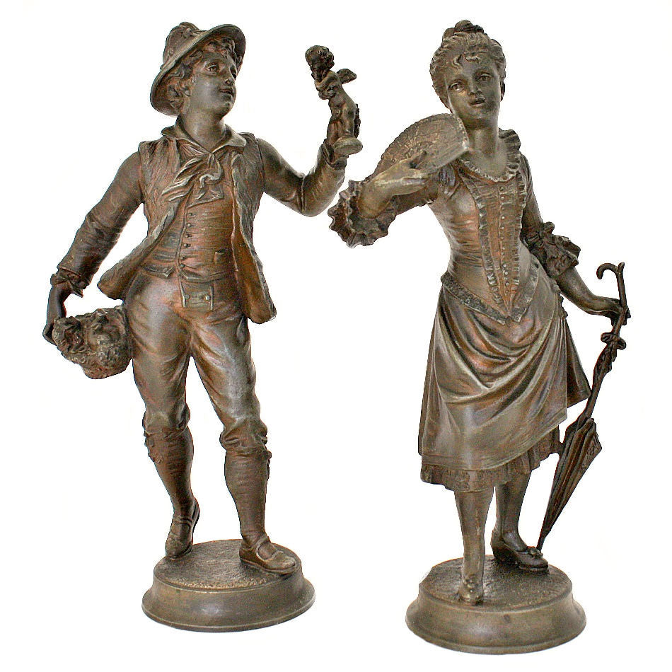 A pair of antique French bronze sculptures, depicting a peddler and a young woman with a fan, placed on separate bases.