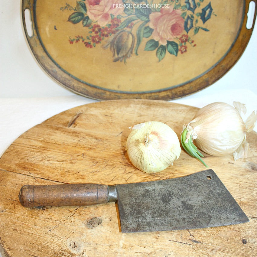 An antique French chef's cleaver with a wooden handle and a blade, displayed on a wooden surface with a floral-patterned plate in the background.