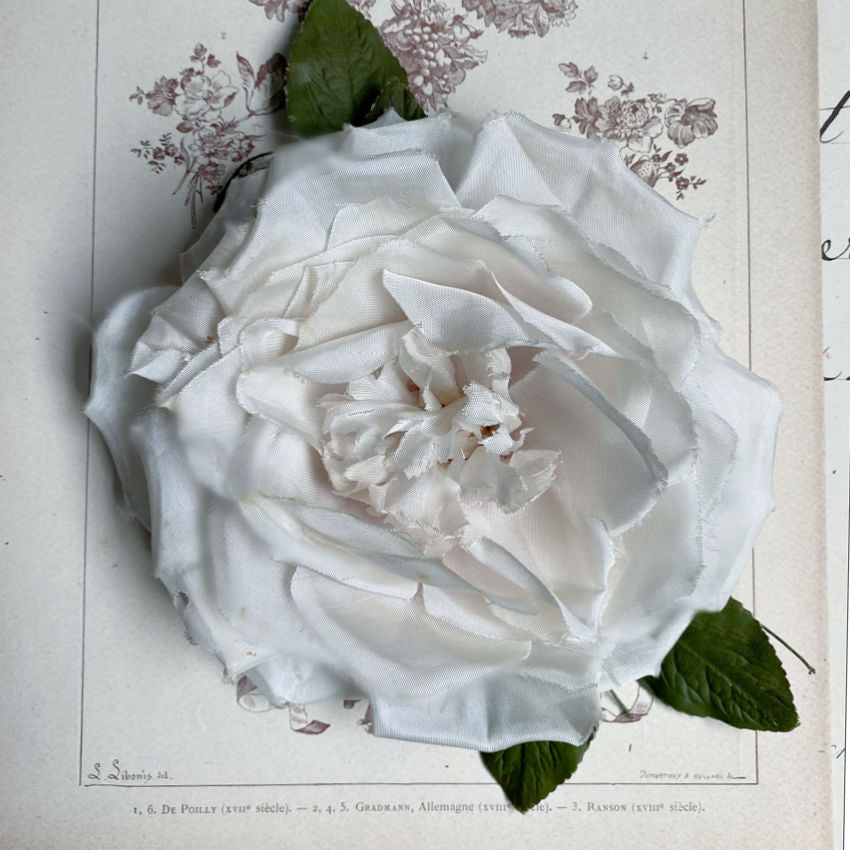 A cream white silk rose peony with green leaves, placed on a surface with written text in the background.