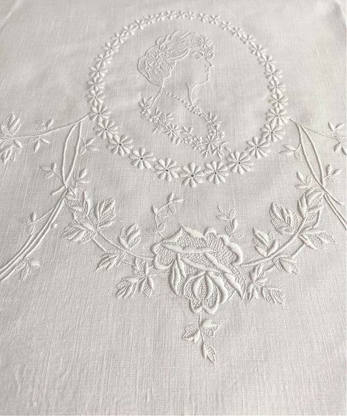 Close-up of white Appenzellembroidered hand towel with a portrait and floral design, showing detailed flower patterns and a monogram.