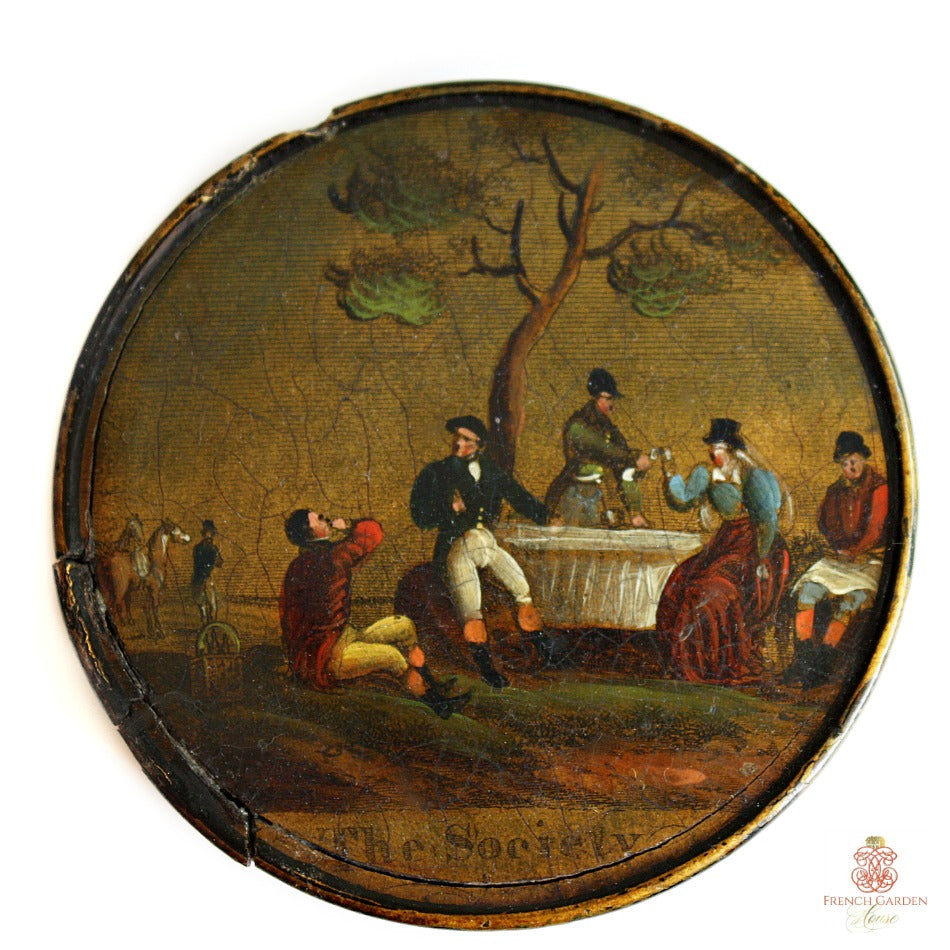An antique circular wooden snuff or patch box with a hand-painted scene of people drinking and socializing outdoors.