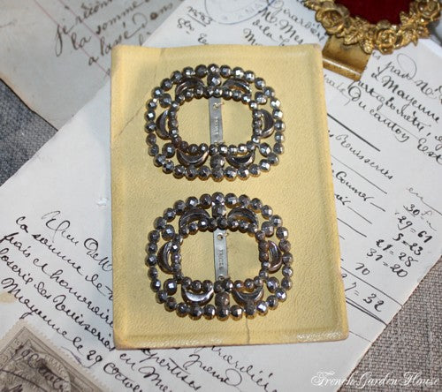 A pair of antique French steel cut shoe buckles displayed on a beige card, with written text in the background.
