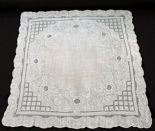 An antique white linen handkerchief with embroidered floral and geometric designs, scalloped edges, and a small border.
