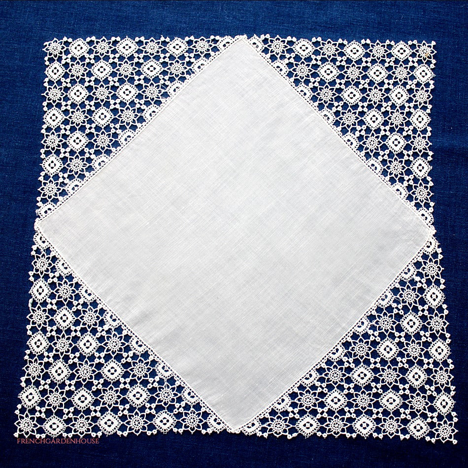 A white linen lawn handkerchief with hand-made needle lace, featuring delicate floral and geometric patterns, displayed on a blue background.