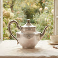 Antique Art Deco silver plated teapot with rounded body, curved handle and long elegant spout on table in front of garden