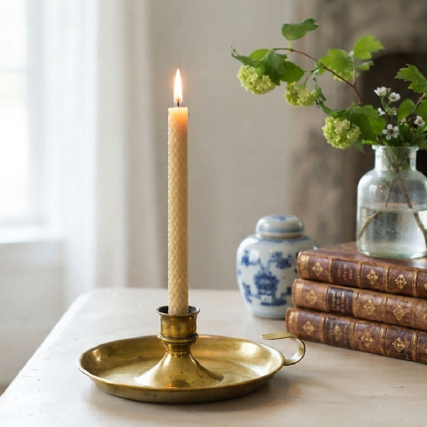 Antique French brass chamberstick with lit candle on a table by a window, beside antique books and a glass vase of green snowball hydangeas and white flowers.