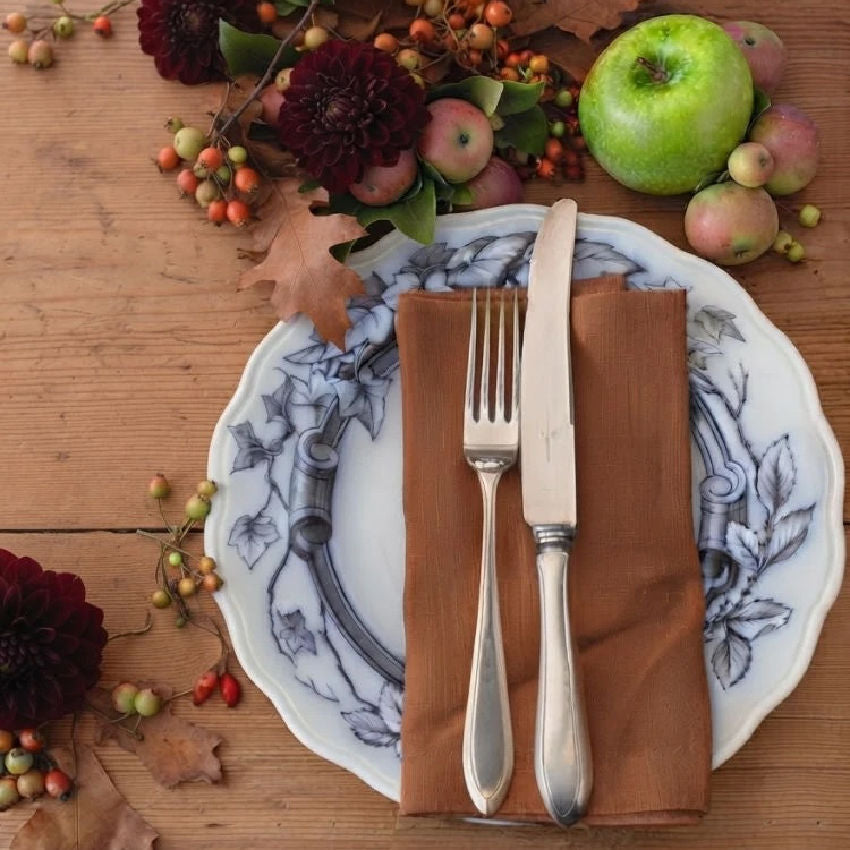 A styled autumn table setting featuring a English Flow blue plate with a ivy and scroll pattern, topped with a brown cloth napkin, a silver fork, and a silver knife. The setting is decorated with dark burgundy dahlias, small red berries, fall leaves, and apples on a rustic wooden table.