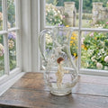 Antique Mary Gregory glass pitcher with hand painted boy and white enamel trees in garden room with view outside of flowers