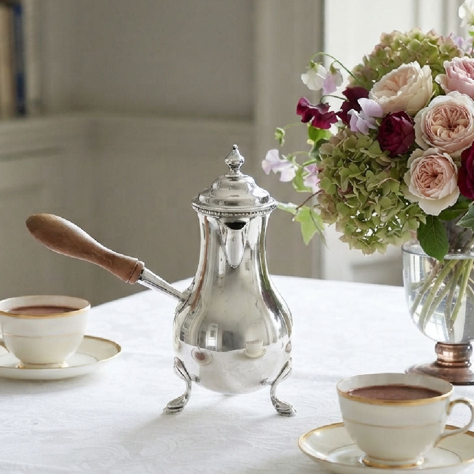 Antique silver plated chocolate pot with turned wood handle on a table with cups of chocolate and a rose and hydrangea bouquet