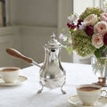 Antique silver plated chocolate pot with turned wood handle on a table with cups of chocolate and a rose and hydrangea bouquet