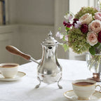 Antique silver plated chocolate pot with turned wood handle on a table with cups of chocolate and a rose and hydrangea bouquet