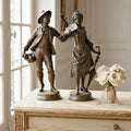 Two bronze classical figurines of a young man and woman standing on a distressed cream-colored wooden cabinet, with a small vase of pale pink roses beside them. French figures