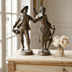 Two bronze classical figurines of a young man and woman standing on a distressed cream-colored wooden cabinet, with a small vase of pale pink roses beside them. French figures
