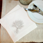 A white linen napkin with embroidered wheat sheaf design, placed on a table with a fork and knife beside it.