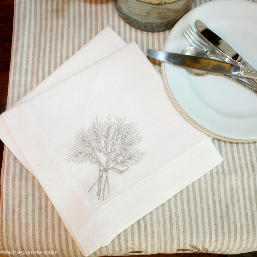 A white linen napkin with embroidered wheat sheaf design, placed on a table with a fork and knife beside it.