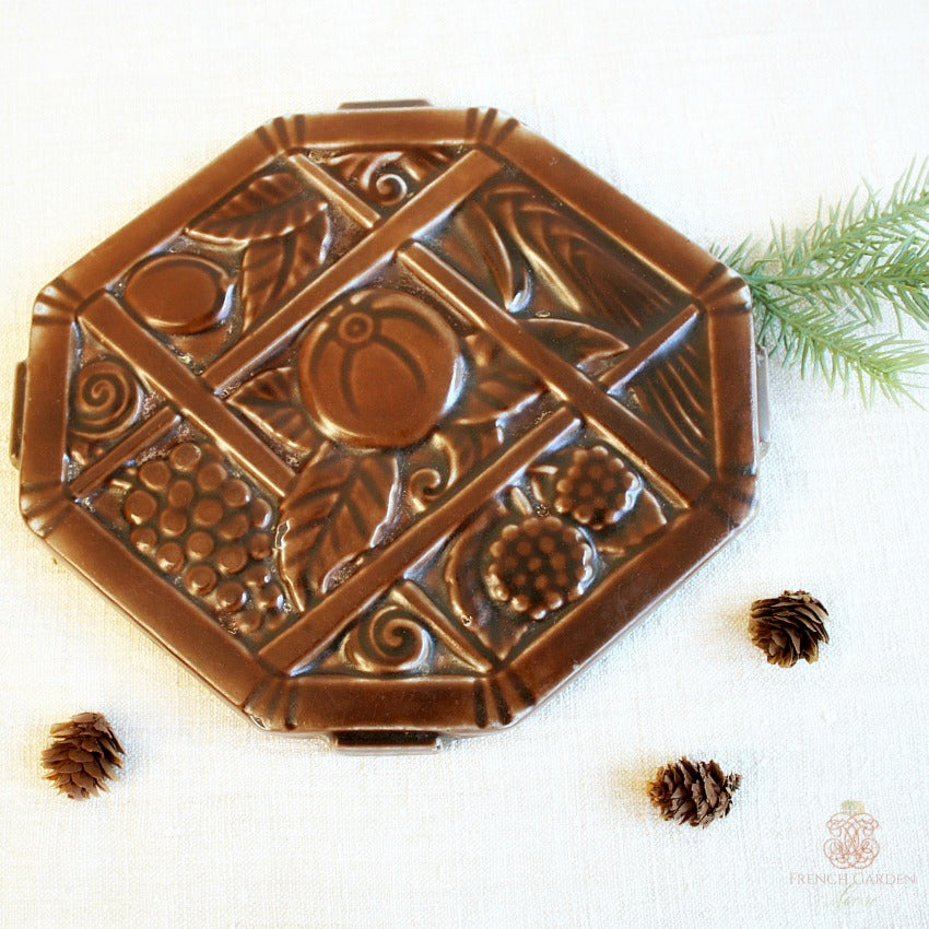 A square-shaped, brown enameled French trivet with Art Deco decorative elements, including geometric patterns and fruit motifs, displayed on a white fabric surface with a few pine cones scattered around.