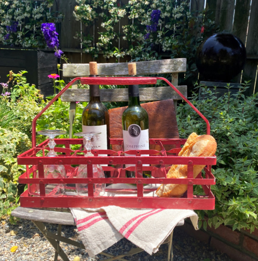 Red metal bottle holder with wine bottles, glasses, and bread on a garden bench.