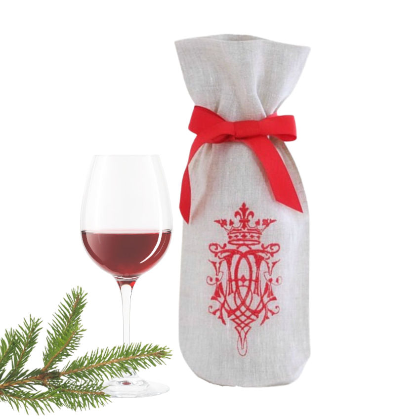 Decorative wine bottle bag with red emblem and bow, next to a glass of red wine and Christmas tree branches on a white background.