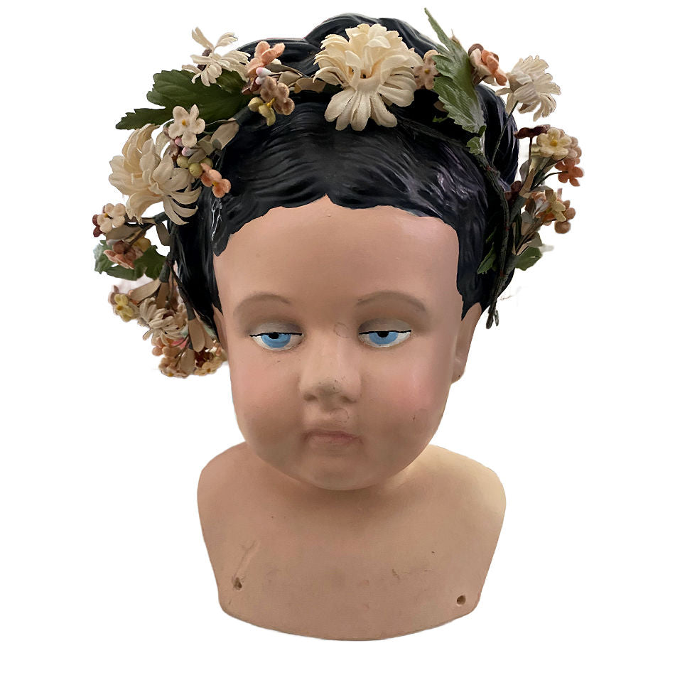 A large pottery doll head featuring an antique millinery floral garland, with a sweet face and blue eyes. The doll has a 'put up' hairdo with a gold 'clip' on the back.