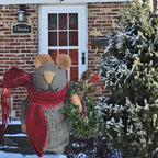 Decorative Felted Christmas mouse figure with a red scarf and wreath in front of a brick house.