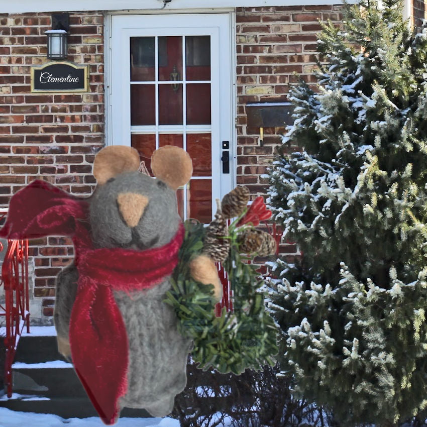 Decorative Felted Christmas mouse figure with a red scarf and wreath in front of a brick house.