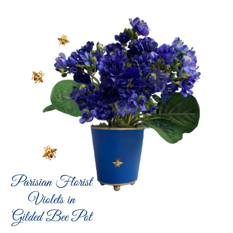 A blue metal tole pot with gilded ball feet and a hand painted rim, containing Parisian blue violets. A small gilded bee is visible on the front of the pot.