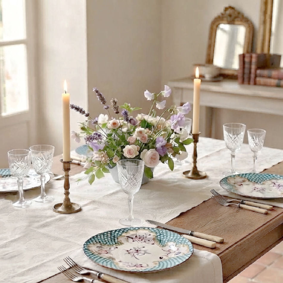 A beautifully styled dining table set for four with floral centerpieces, lit taper candles in brass holders, clear cut-crystal style wine glasses, and matching Antique set of six French hand painted and hand enameled aqua blue plates