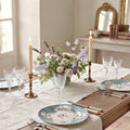 A beautifully styled dining table set for four with floral centerpieces, lit taper candles in brass holders, clear cut-crystal style wine glasses, and matching Antique set of six French hand painted and hand enameled aqua blue plates