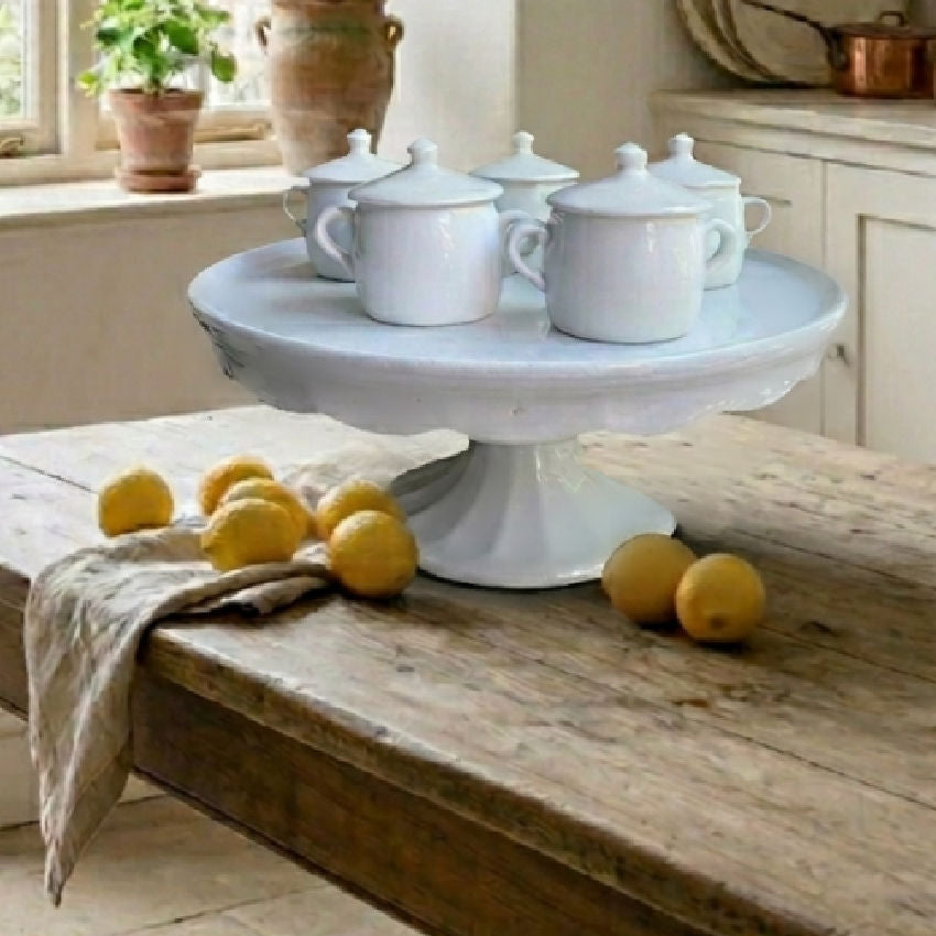 Antique white ironstone cake stand displayed on a rustic French farmhouse table in a sunlit country kitchen with lemons and soft garden light.
