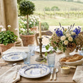 A French country dining table set for four on a stone veranda, featuring antique blue and white Sarreguemines Cérès plates, rustic bread, and a central floral arrangement with blue delphinium and cream roses.