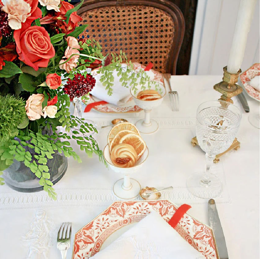 Elegant table setting with floral arrangement, antique plates, and cutlery on a white tablecloth, iwth a milk glass compote at each setting.