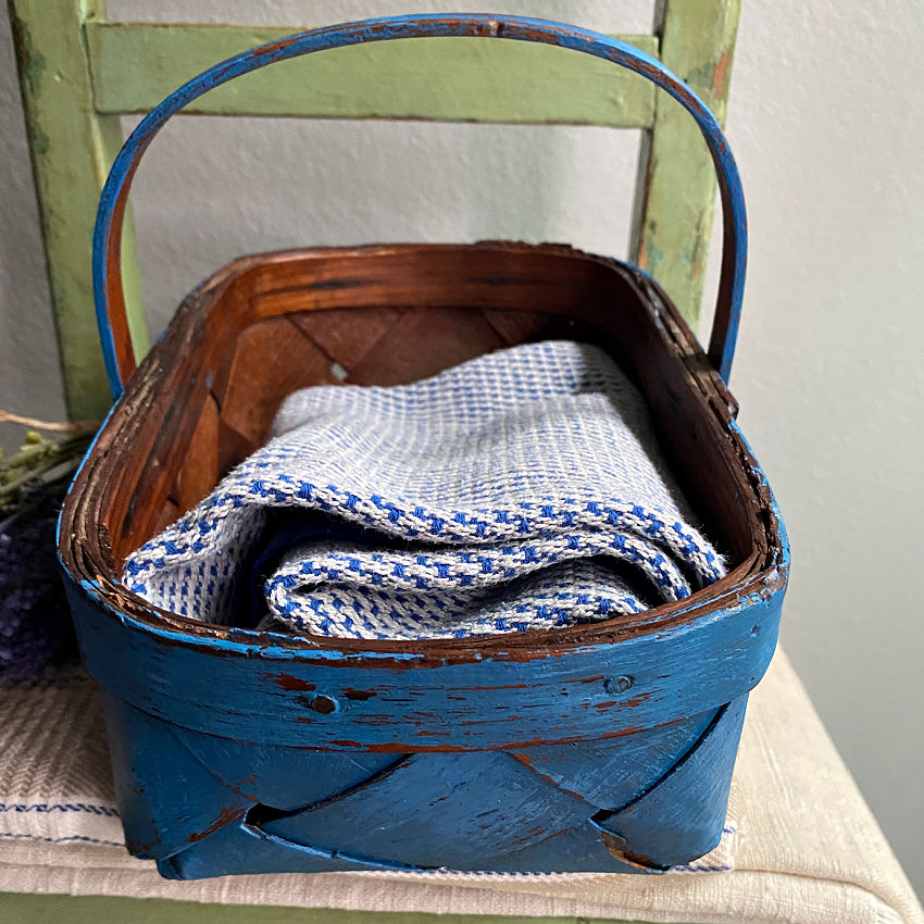 Close-up of the wide splint weave and distressed blue finish on a vintage Normandy basket filled with French blue and white towels.