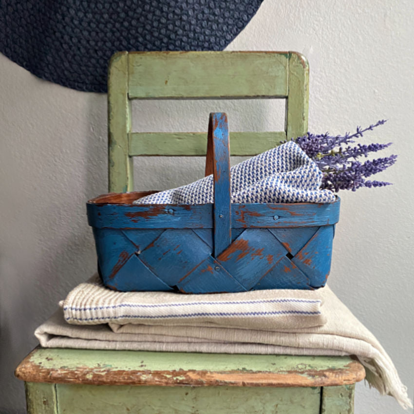 French blue painted  splint weave vintage Normandy basket with a hat, a stack of antique French towels, lavender on an antique chair.
