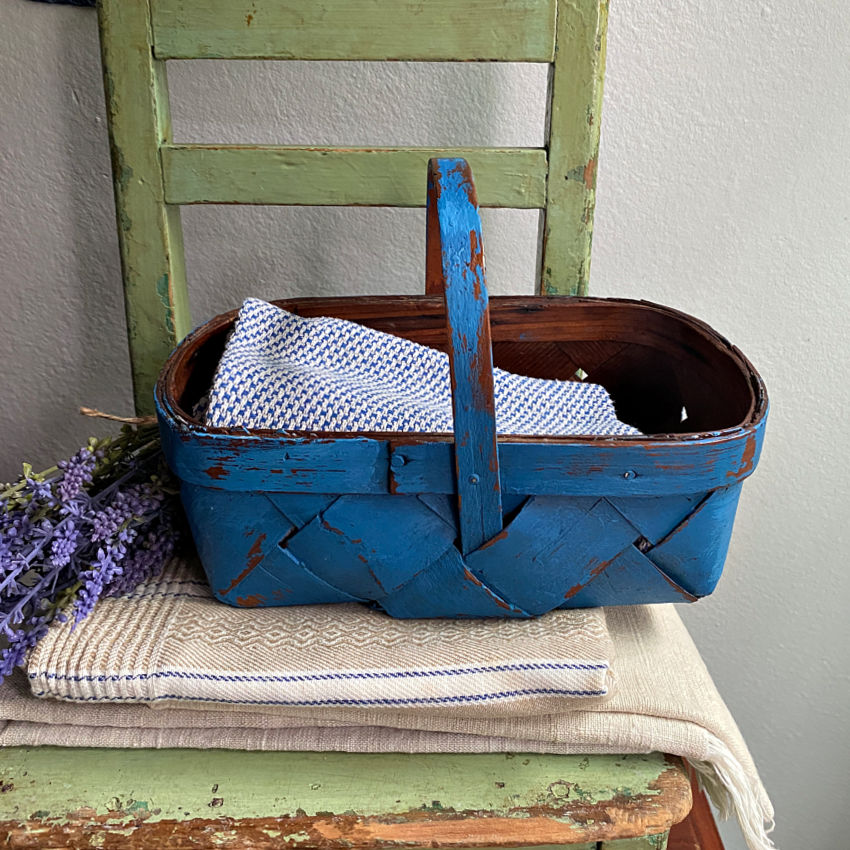 Hand-woven wood splint basket with authentic French blue patina, styled on a stack of antique French towels in blue and cream with lavender.