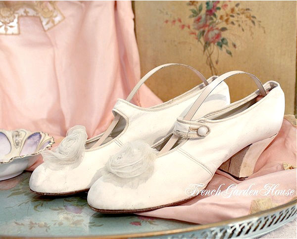A pair of antique Victorian white leather bridal wedding shoes with a French flanged Louis heel and a chiffon flower on the front, displayed next to a pair of pink spanners and a floral embellishment.