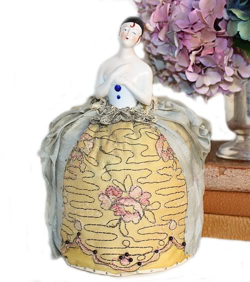 Antique Pierette half doll pin cushion, elegantly gowned, with a golden silk skirt, hand embroidered gold French bullion thread embroidery, pink roses on net, and a sheer French blue bustle. The doll has a white bodice with two painted blue pompoms and hand-painted face with tendrils of hair.